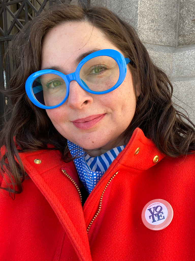 A person with long dark hair is wearing large blue glasses and a bright red coat. They have a VOTE button pinned to their coat. The background features a stone wall with ornate metalwork.
