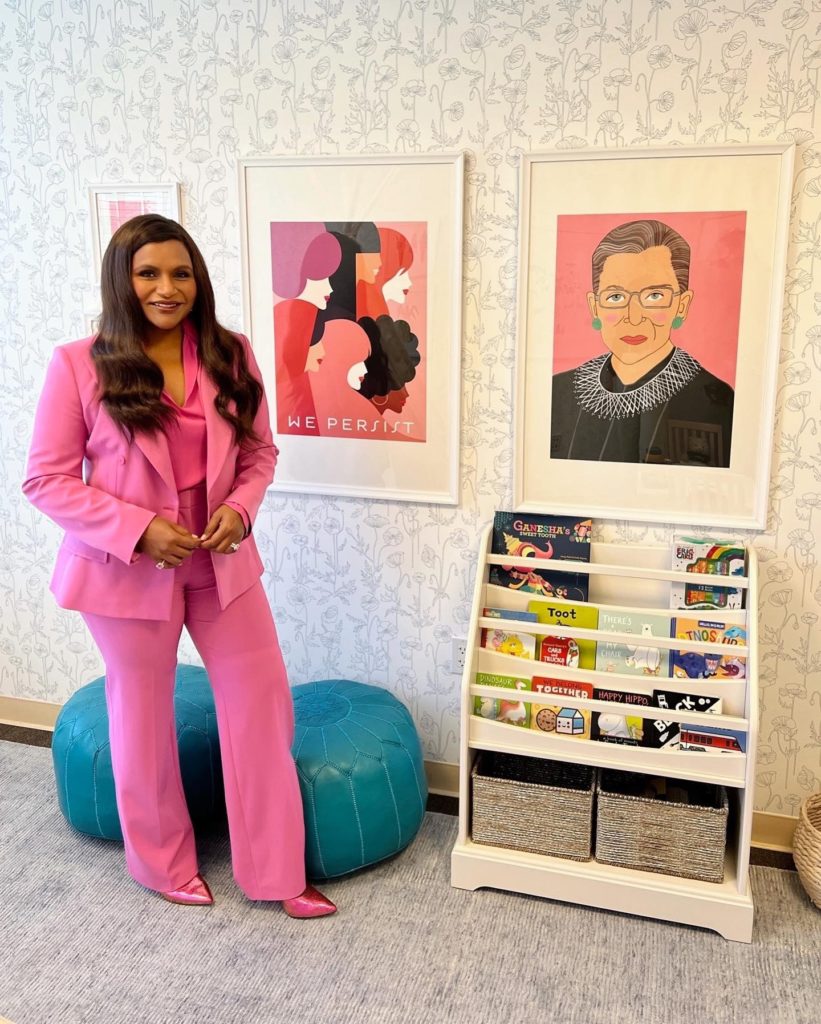 Mindy Kaling, a person in a pink suit stands next to a bookshelf filled with childrens books. Two framed artworks are on the wall: one with abstract faces and We Persist, and another portrait of a woman wearing glasses and a lace collar.